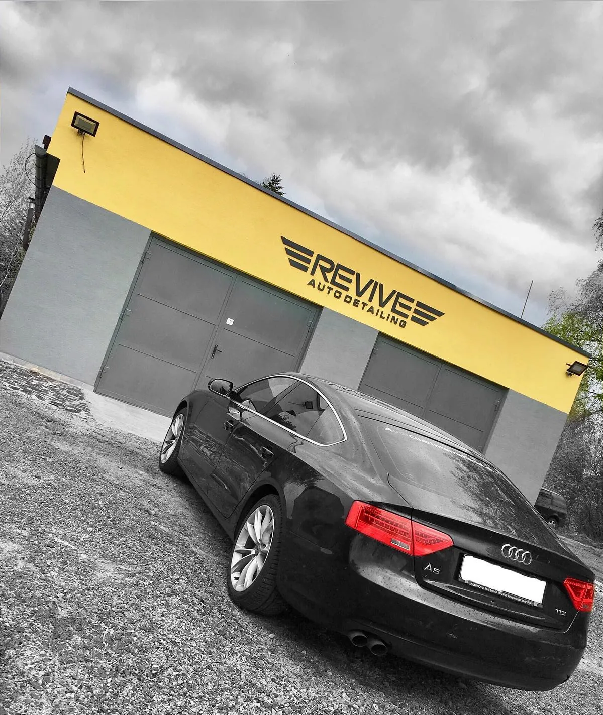 New yellow-and-black studio building with black REVIVE lettering and a black Audi A5 on gravel, photo from 23 May 2019 (black-and-white edit with yellow)