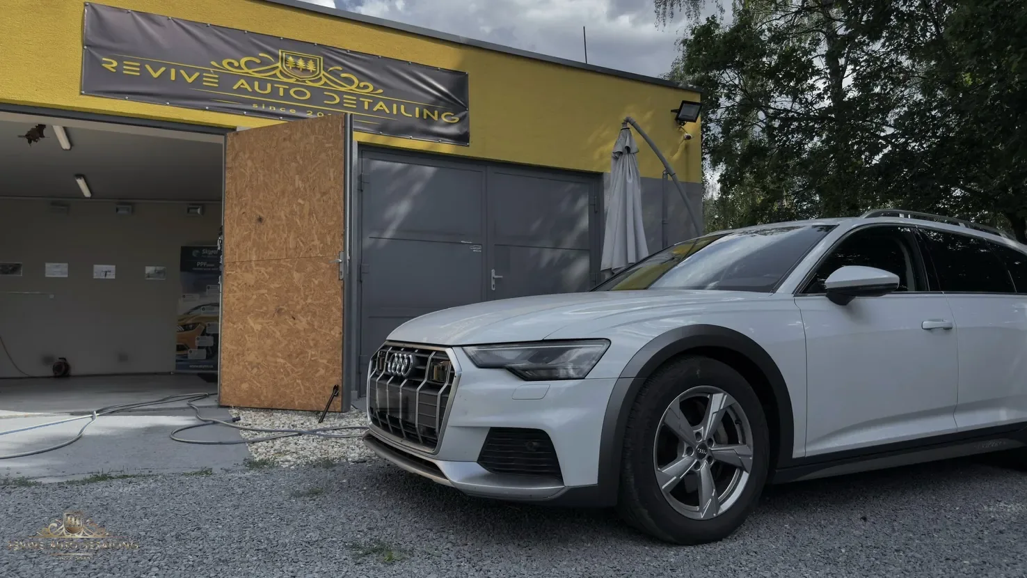 Open studio doors beneath a black banner with a golden crest, with a white Audi A6 allroad on gravel in front